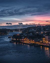 Illuminated city by sea against sky at sunset