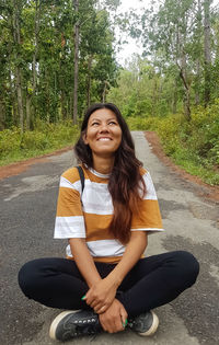 Portrait of smiling young woman sitting on road against trees