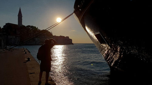 Silhouette woman on illuminated city by sea against sky during sunset