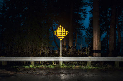Illuminated information sign by trees against sky at night