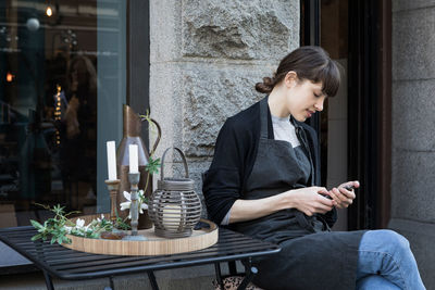 Young female owner using smart phone while sitting by table against store