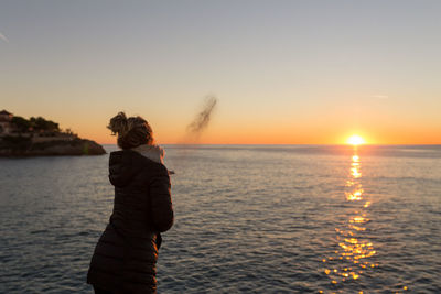 Rear view of woman standing on sea against sky during sunset