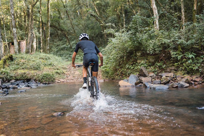 Back view of anonymous male bicyclist riding bike in river with foam during trip in woods