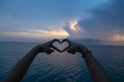 Close-up of hand making heart shape against sea during sunset