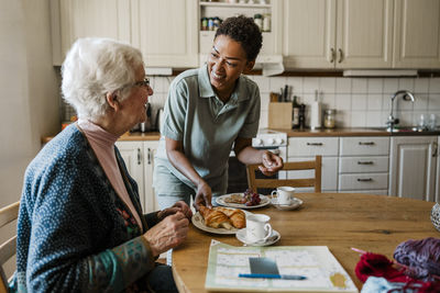 Happy female nurse serving croissant to senior woman sitting at table in kitchen during house call