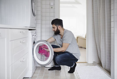 Side view of man crouching while loading clothes in washing machine at home