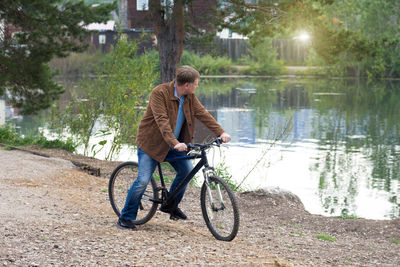 An adult man in a corduroy brown jacket rides a bicycle in a park along the river.