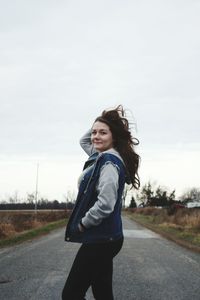 Side view portrait of smiling young woman standing on road against sky