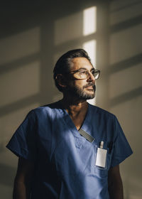 Mature male physician looking away while standing against wall at hospital
