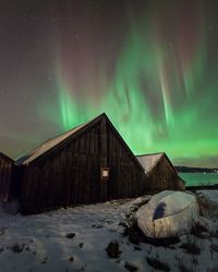 Wooden houses by snowcapped field against aurora borealis in sky