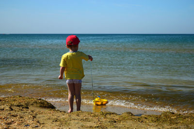 Rear view of boy playing with toy boat while standing at beach against clear sky