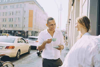 Smiling businessman drinking coffee with female coworker on sidewalk cafe in city
