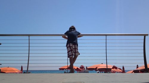 Rear view of boy standing by railing by sea against clear sky