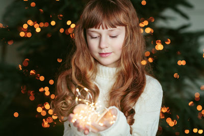 Happy young girl sitting by the christmas tree with garland