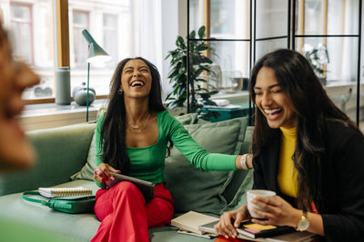 Cheerful businesswoman laughing while sitting with female colleague on sofa at office