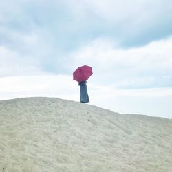 Rear view of woman with umbrella standing on sand at beach against cloudy sky