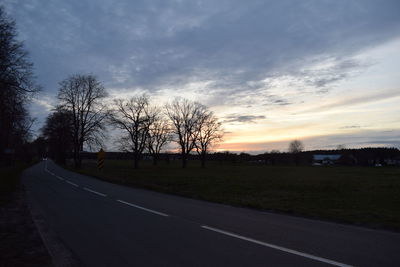 Road by bare trees on field against sky at sunset