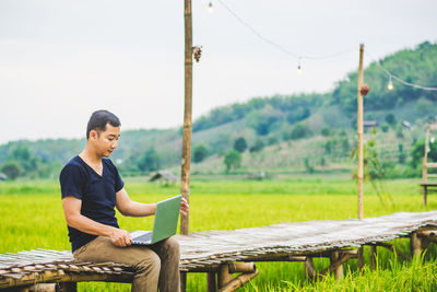 Man using laptop while sitting on boardwalk at rice paddy