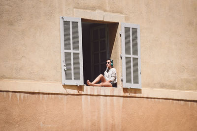 Low angle view of woman hearing music while sitting on window