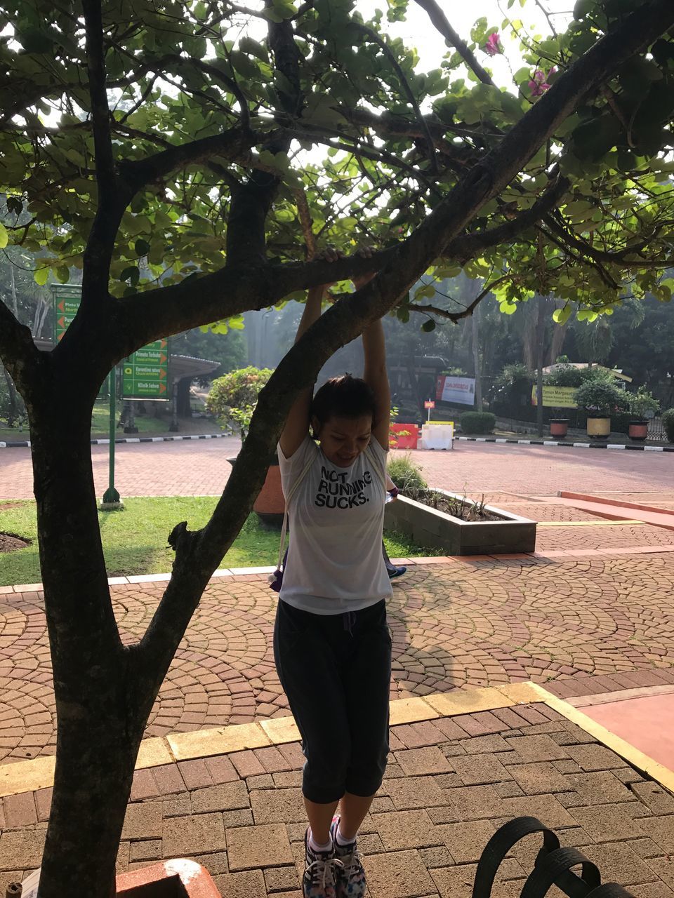 real people, tree, full length, lifestyles, one person, leisure activity, arms raised, front view, young women, young adult, casual clothing, outdoors, park - man made space, day, standing, happiness, growth, looking at camera, built structure, architecture, nature, smiling