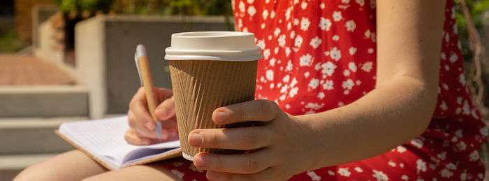 Unrecognizable young woman in red dress drinking coffee from craft paper cup writing gratitude