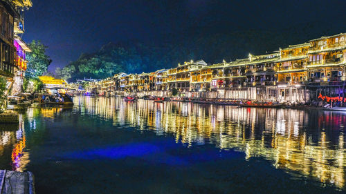 Illuminated buildings by lake against sky in city at night