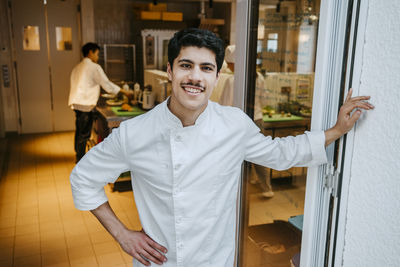 Portrait of happy male chef standing with hand on hip at door in commercial kitchen