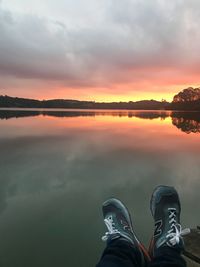 Low section of person by lake against sky during sunset