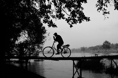 Silhouette man riding bicycle on bridge over lake against sky