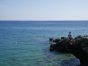 People on rocks by sea against clear sky