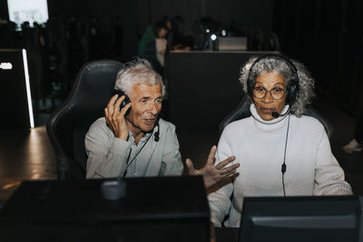 Senior man wearing headset sitting by female friend playing video game on computer at gaming lounge