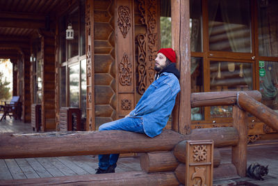 Portrait of a village man with a beard in a red hat stands at a wooden house in autumn