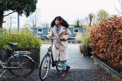 Smiling young african american female with curly dark hair wearing warm coat walking with bicycle while listening to music in headphones on street in autumn day