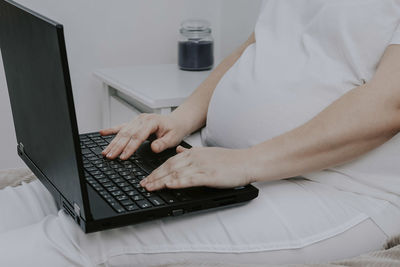 Pregnant woman working on laptop sitting at home in bed, close-up side view.