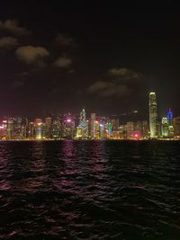 Illuminated buildings by sea against sky at night