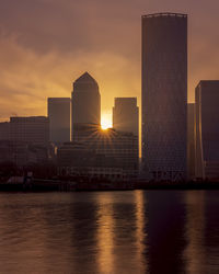 Modern buildings by river against sky during sunset