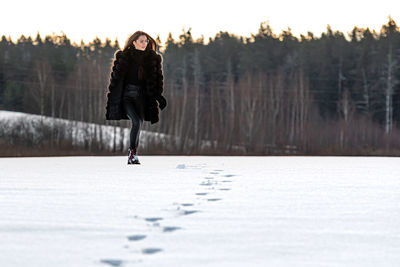 A cheerful woman in a warm fur coat walks across the open field leaving footprint in the snow