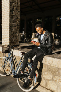 Full body side view of cheerful african american male with backpack sitting with bicycle on paved street near building in city