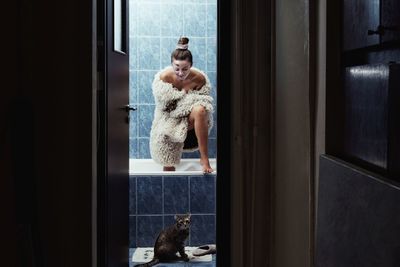 Fashion model wearing costume standing in bathtub with cat at bathroom