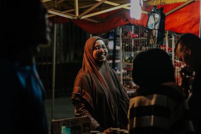 Cheerful female vendor with customers at illuminated market stall during night