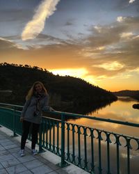 Portrait of smiling woman standing on footbridge over river against cloudy sky during sunset