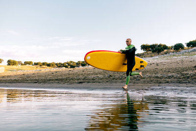 Full body of active sporty male in wetsuit carrying paddle board and entering river water