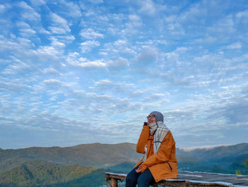 Young woman in warm clothing sitting on observation point against cloudy sky