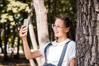 A schoolgirl in glasses takes a selfie on a smartphone near a tree in the park.