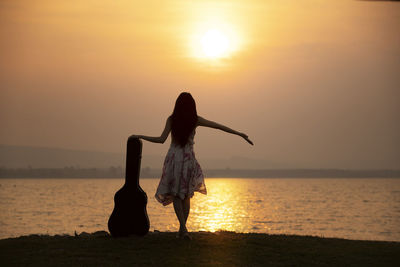 Rear view of woman with guitar container at beach against sky during sunset