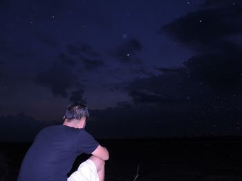 Rear view of man sitting on rock against sky at night