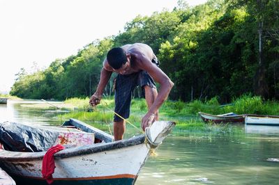 Shirtless fisherman tying rope in boat on river