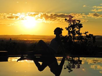 Silhouette man sitting by lake against sky during sunset