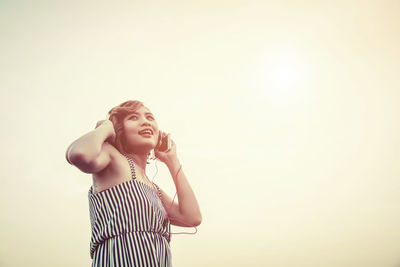 Woman listening music while standing against clear sky during sunset