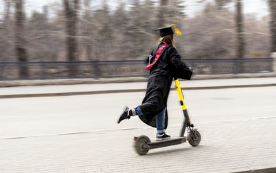 Young woman in black graduation gown and graduation cap riding an electric scooter after graduation 